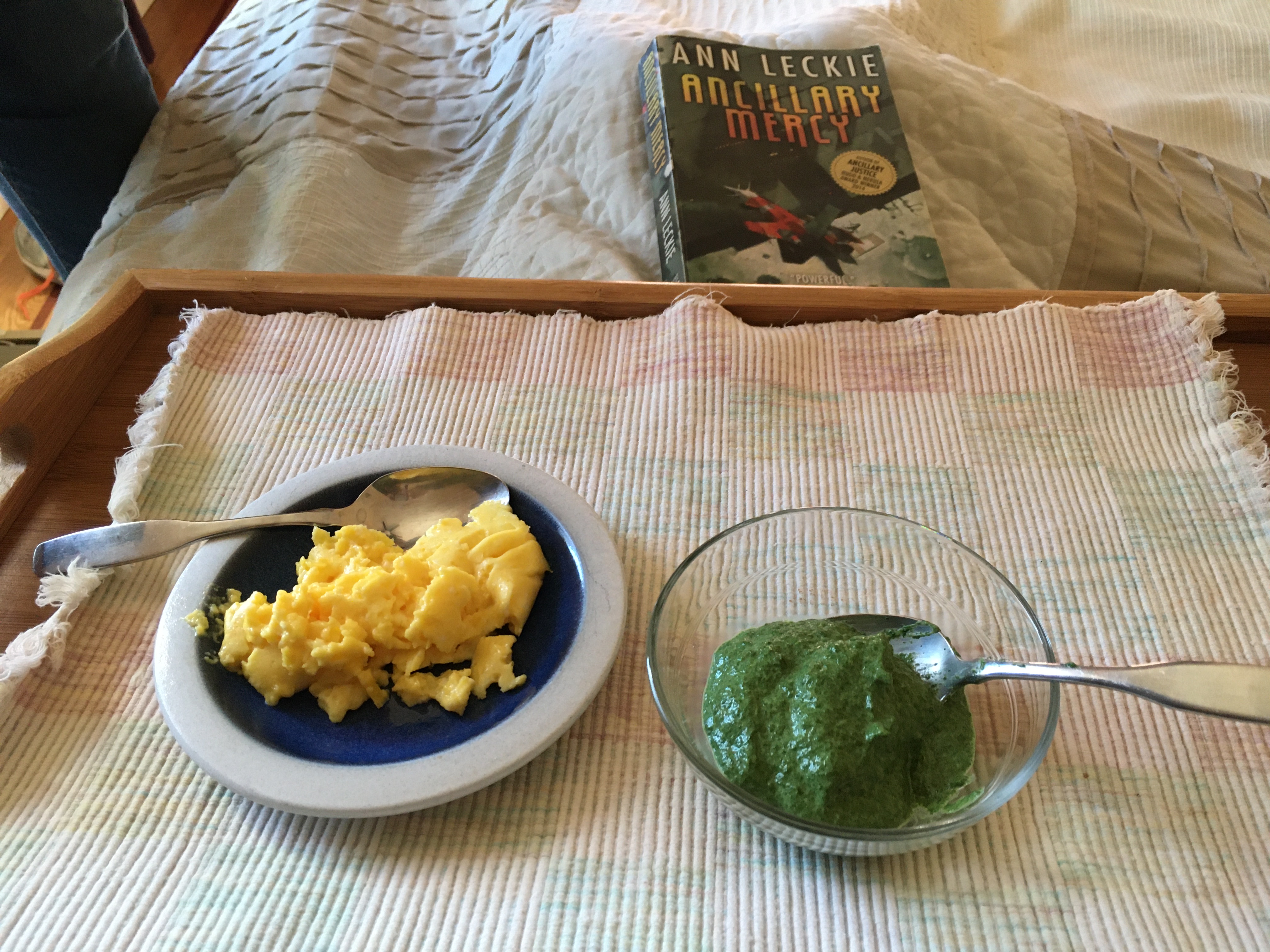 A food tray with two items: small plate of scrambled eggs, small bowl of creamed spinach. A book Ancillary Justice is in the background.