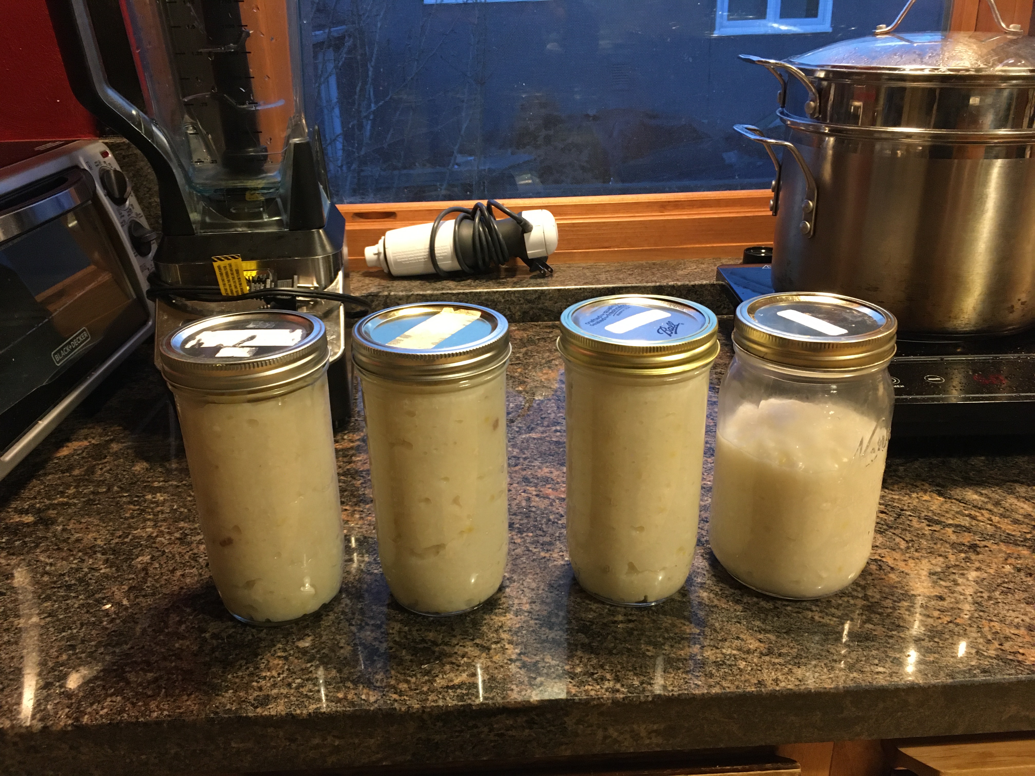 4 mason jars filled with white liquids on a kitchen counter, with a blender, stick blender, and pot behind them.